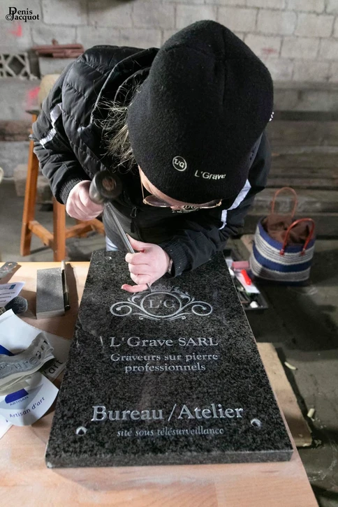 Deux artisans graveurs dans leur atelier de pierre, l'une manipulant un outil de gravure traditionnelle sur une dalle de granit avec concentration, l'autre observant ou préparant un élément en marbre. L'atelier doit montrer des outils professionnels, de la pierre, des matériaux de gravure, et refléter un environnement de travail artisanal authentique et passionné. L'image doit capturer la complicité professionnelle et l'expertise technique du duo.