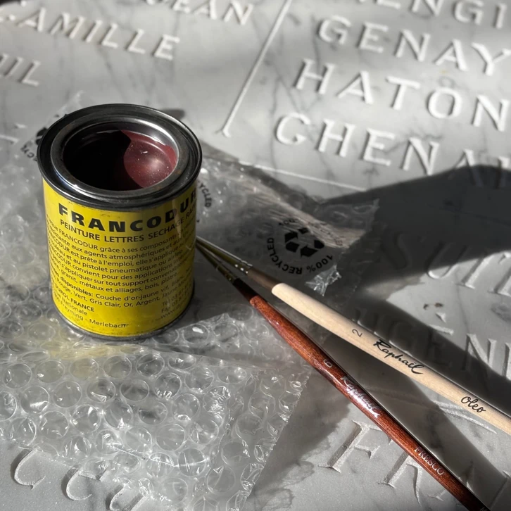 Atelier intérieur de gravure sur pierre montrant un artisan graveur au travail devant une machine laser haute performance en fonctionnement, en train de graver une plaque en granit. L'atelier doit montrer du matériel de gravure professionnel, des pièces de pierre (granit, marbre) en attente de travail ou finalisées, et refléter l'environnement d'un atelier d'artisan graveur funéraire moderne avec technologie laser. L'ambiance doit être sérieuse et professionnelle.